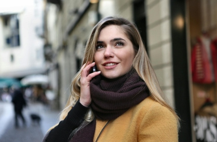 Mujer con abrigo mostaza y bufanda hablando por teléfono en la calle